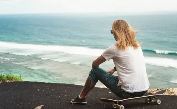 Jovem homem skatista loira sentado na frente da vista incrível do oceano — Fotografia de Stock