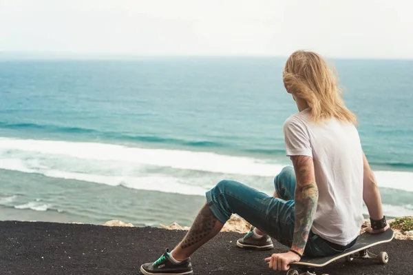 Jovem homem skatista loira sentado na frente da vista incrível do oceano — Fotografia de Stock