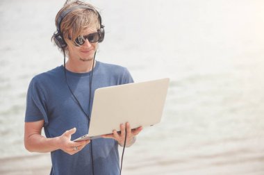 Laptop ve kulaklık ile plajda çalışan yakışıklı sarışın adam