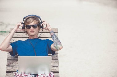 Laptop ve kulaklık ile plajda çalışan yakışıklı sarışın adam