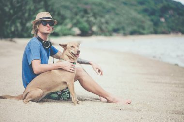Mavi tişört, şapka ve güneş gözlüğü ile köpek sahilde oturup, giyen genç yakışıklı adam