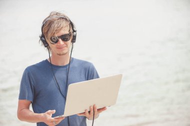 Laptop ve kulaklık ile plajda çalışan yakışıklı sarışın adam