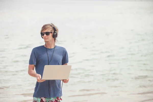 Bonito homem loiro trabalhando com laptop e fones de ouvido na praia — Fotografia de Stock