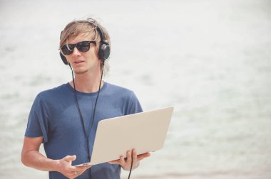 Laptop ve kulaklık ile plajda çalışan yakışıklı sarışın adam