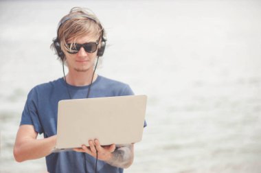 Laptop ve kulaklık ile plajda çalışan yakışıklı sarışın adam