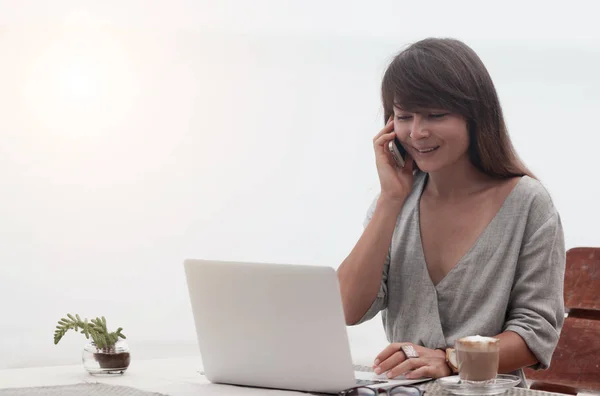 Junges schönes Mädchen sitzt in einem Café mit einem Laptop und spricht auf der Pnone — Stockfoto