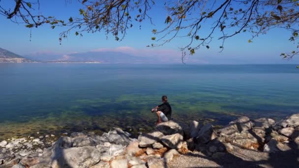 Чоловік сидить на камені на узбережжі озера — стокове відео