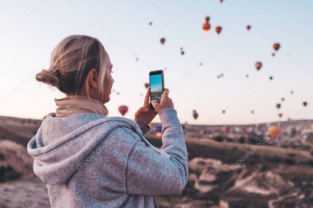 Woman taking photo on beautiful landscape and balloons in Cappadocia with mobile camera, sunrise time.