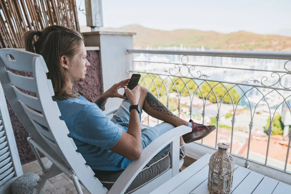 Mężczyzna siedzi na tarasie w hotelu i używa telefonu komórkowego — Zdjęcie stockowe