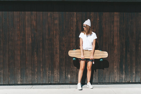 Attractive woman in sunglasses and hat standing at the wall with longboard