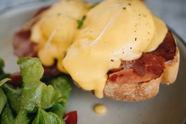 Benedict yumurtası, domuz pastırması ve beyaz tabakta salata mermer arka planda.