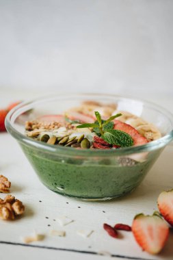 Muz ve spirulinalı meyve suyu kasesi, badem gevreği, chia tohumu, kabak çekirdeği ve beyaz arka planda çilek..