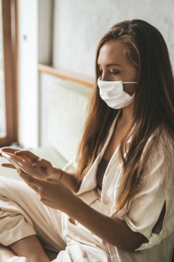 Maskeli genç bir kadın evdeki kanepede oturan telefonu kullanıyor.