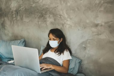 Maskeli kadın dizüstü bilgisayarla yatakta yatıyor. Boşluğu kopyala.