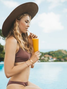 Yüzme havuzunda oturmuş mango içen, yaz şapkalı, gülümseyen sarışın kadın.
