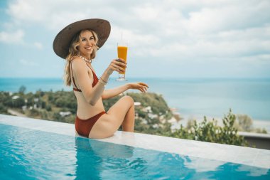 Yüzme havuzunda oturmuş mango içen, yaz şapkalı, gülümseyen sarışın kadın.