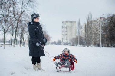 Büyükanne ve torunu bir kış gün parkta halletme. renkli snowsuits bir kızak yolculuğu zevk üzerinde yürümeye başlayan çocuk