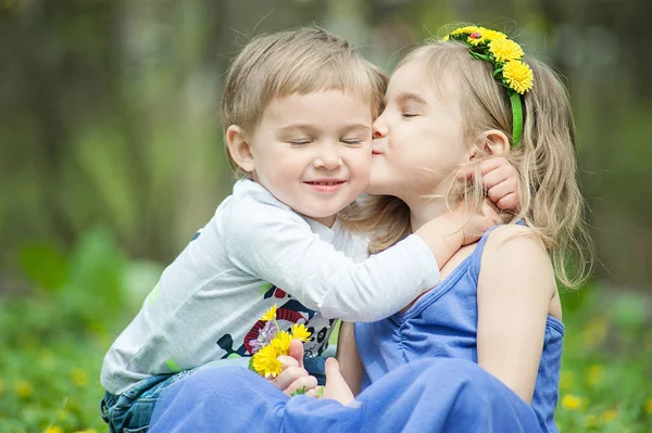 Brother and sister sit on the grass. Children's games, leisure. Two children are sitting on green meadow and smile. Boy and girl kiss on sunny day.