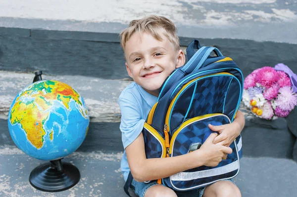 First-grader is sitting with a backpack on the doorstep of the school. bouquet and globe. the first call. end of the school year — Stock Photo, Image