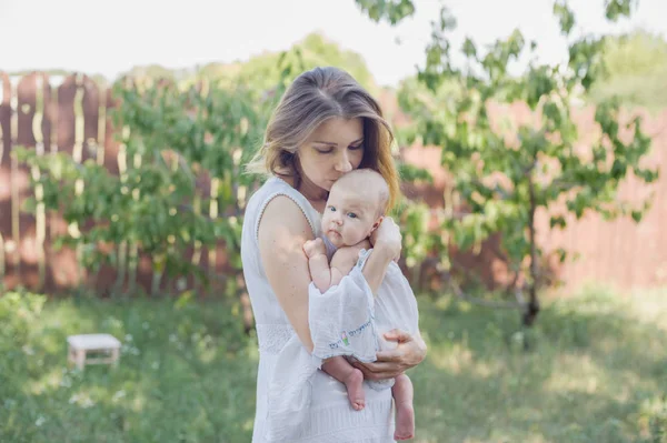 Eine schöne junge Mutter mit einem Baby auf dem Arm steht im Garten. — Stockfoto