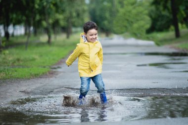 Su birikintisine oynarken küçük çocuk yağmurluk ve lastik çizmeler