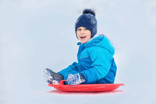 Ребенок катится по снежному холму. Мальчик скользит по снежному холму зимой. Дети играют на улице. Концепция зимних развлечений — стоковое фото