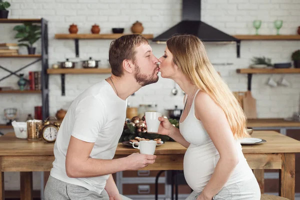 Έγκυος γυναίκα και ο σύζυγος Έχοντας πρωινό στην κουζίνα πίνουν καφέ ή φρέσκο χυμό και φιλιά. — Φωτογραφία Αρχείου