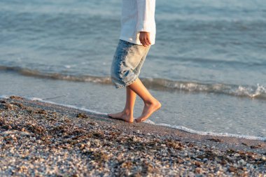 Küçük çocuk sahilde yürüyor, mavi denizde su sıçrıyor.