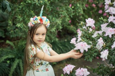 Fantasy little girl in rainbow unicorn horn headband posing near azalea flowers in park 