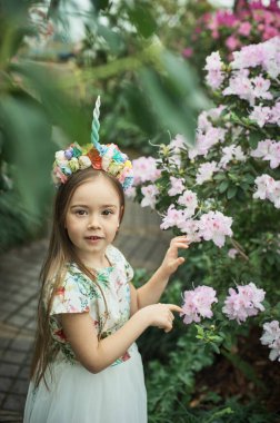 Fantasy little girl in rainbow unicorn horn headband posing near azalea flowers in park 