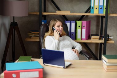 Ofis masasında laptopla çalışırken akıllı telefondan konuşan hamile bir kadın. 