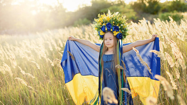 милая девочка в цветочном венке с лентами, с голубым и желтым украинским флагом и прогулкой по летнему лугу, концепция Дня Независимости Украины, День Государственного флага Украины и День Конституции, Патриотический праздник
  