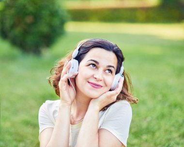 Kulaklıkla müzik dinleyen ve parkta çimlerin üzerinde oturan mutlu hamile bir kadın. Hamilelik ve Sağlık Hizmetleri kavramı.