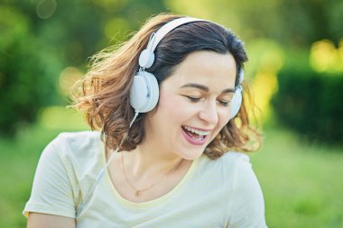 Kulaklıkla müzik dinleyen ve parkta çimlerin üzerinde oturan mutlu hamile bir kadın. Hamilelik ve Sağlık Hizmetleri kavramı.