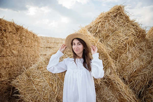 Young girl wears summer white dress near hay bale in field. Beautiful girl on farm land. Wheat yellow golden harvest in autumn. Countryside natural landscape.