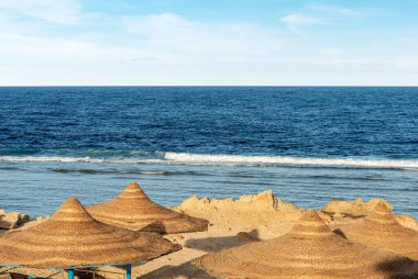 Marsa Alam, Mısır, Afrika yakınlarında saman şemsiyeli Kızıldeniz plajı. Deniz dalgaları mercan resiflerine çarpar.