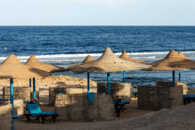 Marsa Alam, Mısır, Afrika yakınlarında saman şemsiyeli ve şezlonglu Kızıldeniz plajı. Deniz dalgaları mercan resiflerine çarpar.