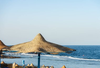 Marsa Alam, Mısır, Afrika yakınlarındaki Kızıldeniz plajında saman şemsiyeler. Deniz dalgaları mercan resiflerine çarpar.