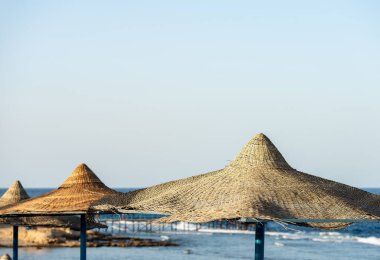 Marsa Alam, Mısır, Afrika yakınlarındaki Kızıldeniz plajında saman şemsiyeler. Arka planda mercan resifinde dalgalar olan deniz.