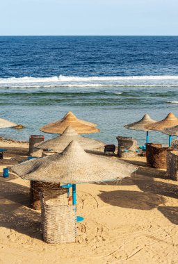 Marsa Alam, Mısır, Afrika yakınlarında saman şemsiyeli Kızıldeniz plajı. Deniz dalgaları mercan resiflerine çarpar.
