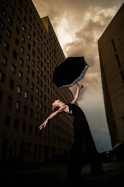 Ballerina with umbrella on city street on background sky and bui