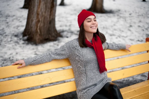 Girl in red hat and scarf is resting on bench in winter park.