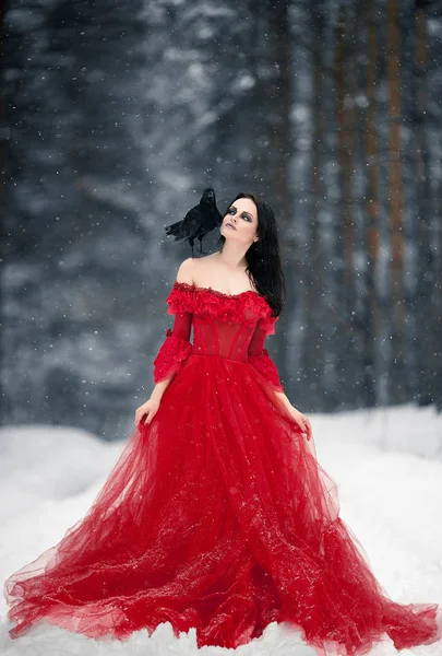 Woman witch in red dress and with raven on her shoulder in snowy forest