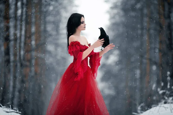 Woman witch in red dress and with raven in her hands in snowy forest