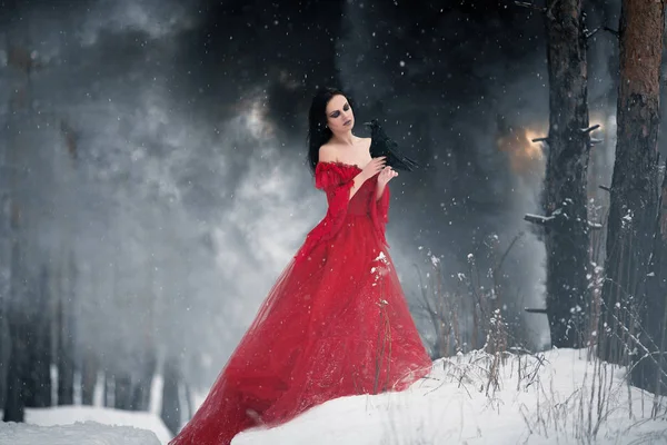 Woman witch in red dress and with raven in her hands in snowy forest. Around can be seen fire and smoke.