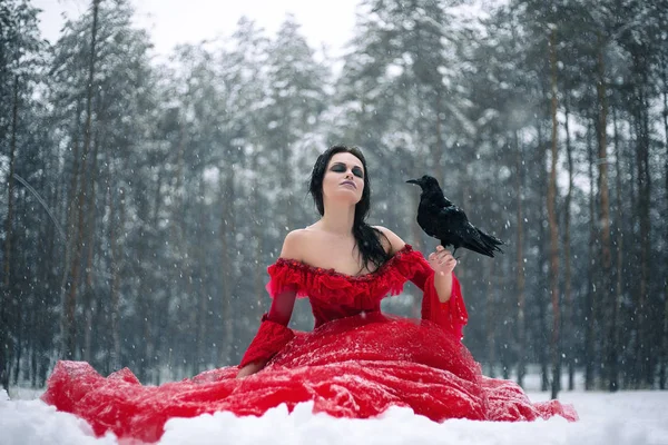 Woman witch in red dress with raven in her hand sits on snow in forest