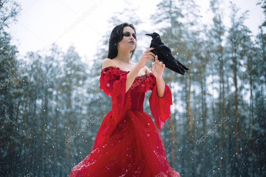 Woman witch in red dress and with raven in her hands in snowy forest
