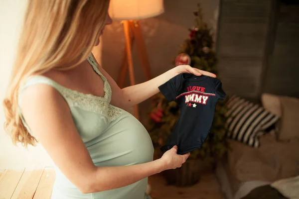 Mujer embarazada se para y sostiene la camisa para su futuro bebé . — Foto de Stock