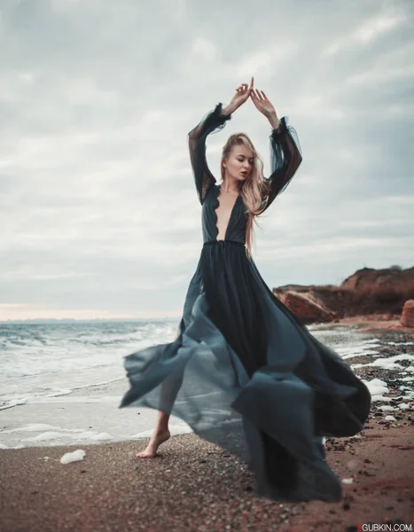 Junge Frau im langen transparenten schwarzen Kleid zwischen Strand und Meer. — Stockfoto
