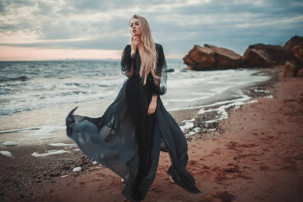 Junge Frau im langen transparenten schwarzen Kleid zwischen Strand und Meer. — Stockfoto